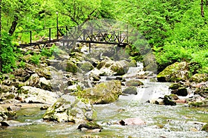 A metal bridge across the rocky bed of a stormy stream flowing down from the mountains through the morning summer forest