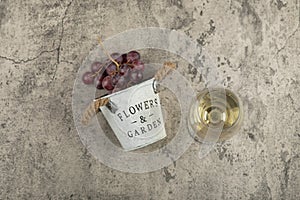 Metal bucket of red fresh grapes and glass of white wine on marble surface