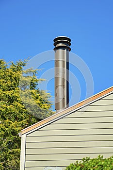 Metal chimney vent on top of house or home with beige wooden horizontal pannels and back and front yard trees with blue