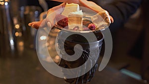 Metal cup with beverage on the bar. Woman put on top of bowl metal sauser with pieces of cheese, dry roses and raspberry