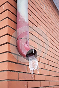 Metal downpipe with frozen stream of water at brick house wall outdoors in winter season.