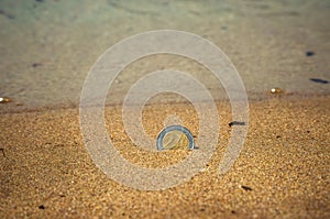 A metal euro coin in the sand as a background. Shallow depth of field. In the background, a washed-out sea with glare from the sun
