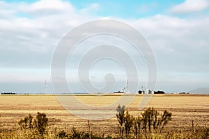 Metal grain elevators on the horizon viewed across harvested wheat field  with fence in foreground under big sky