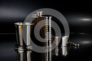 A metal pocket flask for alcoholic beverages with a folding metal cup, shot against a dark background