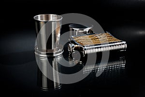 A metal pocket flask for alcoholic beverages with a folding metal cup, shot against a dark background