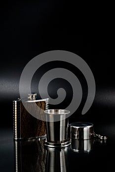 A metal pocket flask for alcoholic beverages with a folding metal cup, shot against a dark background