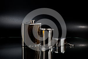 A metal pocket flask for alcoholic beverages with a folding metal cup, shot against a dark background