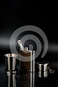 A metal pocket flask for alcoholic beverages with a folding metal cup, shot against a dark background