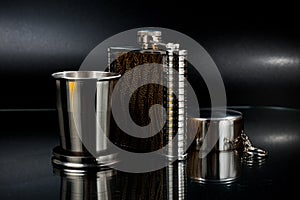 A metal pocket flask for alcoholic beverages with a folding metal cup, shot against a dark background