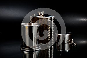 A metal pocket flask for alcoholic beverages with a folding metal cup, shot against a dark background