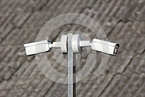 Metal pole with two white surveillance cameras mounted on each side with roof of abandoned industrial complex building in back