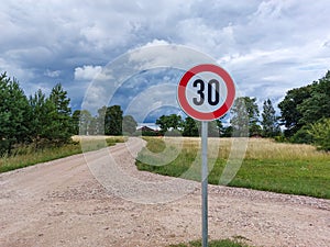 Metal road or traffic sign with number 30 in a red circle indicating new speed limit a gravel road surrounded with countryside