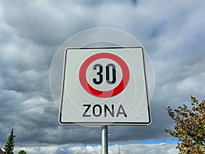 Metal road or traffic sign with number 30 in a red circle, and zone, indicating speed limit against blue sky and clouds background