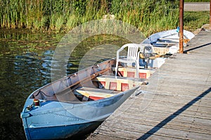 Metal row boats - one with plastic chair in it- pulled up to pier with mossy weedy pond bank behind