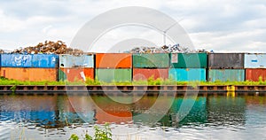 Metal scrap dump, with rusty metal parts on colorful, old truck containers, near water canal, river. Wind power windmills on backg