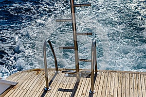 Metal staircase for descent into the water and wave trace with white foam on a water surface behind of fast moving yacht