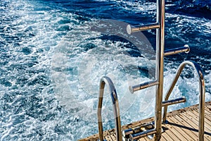 Metal staircase for descent into the water and wave trace with white foam on a water surface behind of fast moving yacht