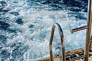 Metal staircase for descent into the water and wave trace with white foam on a water surface behind of fast moving yacht