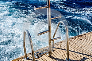 Metal staircase for descent into the water and wave trace with white foam on a water surface behind of fast moving yacht