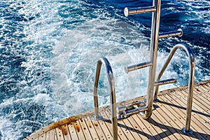 Metal staircase for descent into the water and wave trace with white foam on a water surface behind of fast moving yacht