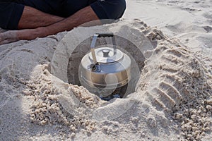 Metal teapot on camping kettle fire burner in a hole dug in the sand on a beach. Travel camping outdoor portable hot water