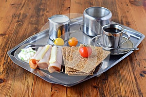 Metal tray with crispbread, cheese, cottage cheese, ham, cherry tomato, cup of coffee, sugar jar, creme jar on a wooden