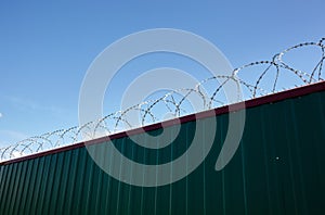 Metal wall with barbed wire against the blue sky background.