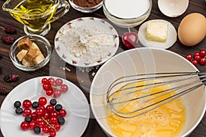 Metal whisk in white bowl with beaten eggs. Berries, flour, butter on table. Dark wooden background