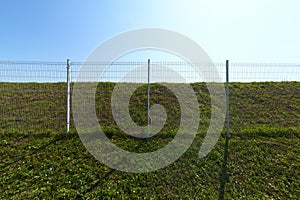 Metal wire industrial grid fence on green grass with blue sky background wide angle shot.