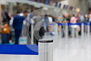 A metallic barrier pole with a blue retractable belt with waiting passengers in a blurred background located in an airport