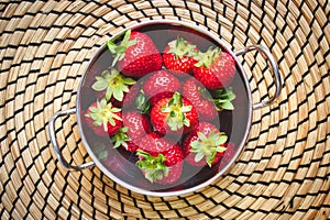 A metallic bowl of red ripe strawberries on a circular straw mat, viewed from above