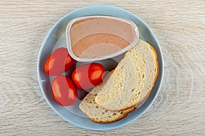 Metallic jar with liver pate, slices bread, tomato in plate on wooden table. Top view
