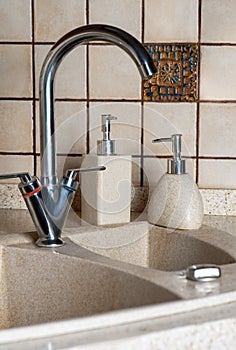 Metallic kitchen faucet, sink from stone and some bottles with soap and cleaner liquids, small ceramic tiles on the wall.