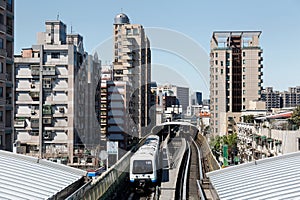 A metro train travels on elevated rails between office towers and residential blocks, arriving at a station of Taipei MRT System