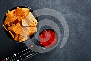Mexican nachos in a black bowl with hot salsa dip sauce and tv remote on black background