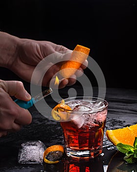 Mezcal Negroni cocktail preparation. Italian aperitivo. Orange peel is burnt in hands.