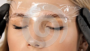Microblading eyebrows work flow in a beauty salon. Cosmetician putting on film on eyebrows.
