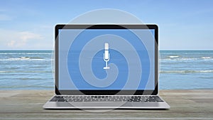 Microphone icon on modern laptop computer screen on wooden table over tropical sea and blue sky with white clouds, Business c