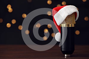 Microphone with Santa hat on wooden table against blurred lights, space for text. Christmas music
