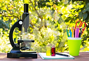 Microscope and stationery on wooden table. Glass flasks with colored liquids Natural green blur background. School concept.