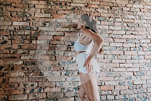 A mid-30s happy woman with long hair natural color poses in her underwear, smiling in front of a brick wall on a