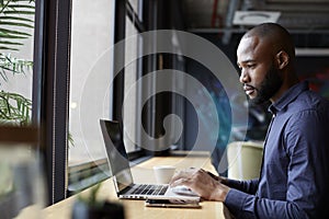 Mid adult black male creative sitting by window in an office social area using a laptop, side view