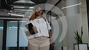 Mid adult black woman in blouse holding digital tablet while walking in office