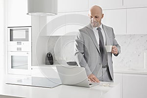 Mid adult businessman having coffee while using laptop in kitchen