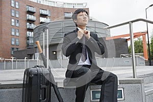 Mid adult businessman with luggage adjusting necktie against buildings