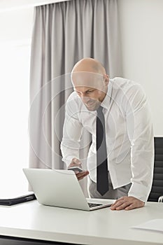Mid adult businessman using call phone and laptop at table in home office
