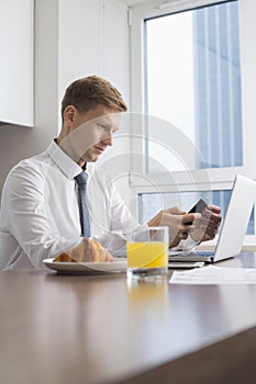 Mid adult businessman using cell phone with laptop on breakfast table