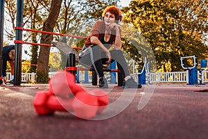 Mid adult Caucasian smiling woman is sitting on playground and listening to music with headphones. Low angle view. Copy