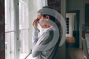 Mid-adult caucasian woman with severe headache standing by the living room window in morning and pressing the forehead with