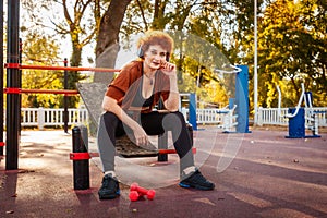 Mid adult Caucasian woman is sitting on playground and listening to music with headphones looking at camera. Autumn park